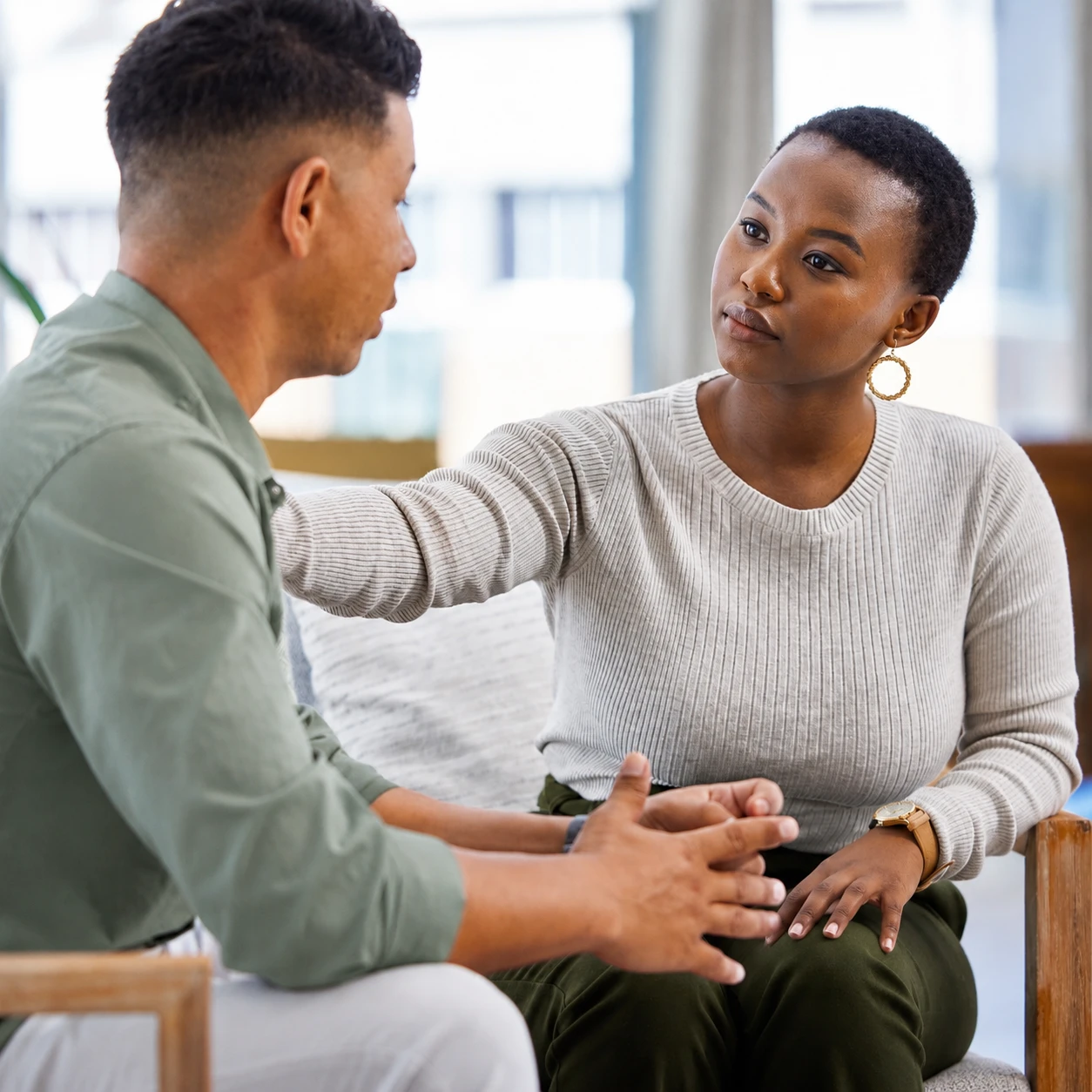 A woman sits beside a man, gently resting her hand on his shoulder as they talk. She looks at him attentively, offering comfort and support during their conversation about communication after stroke. They are seated indoors on a sofa.