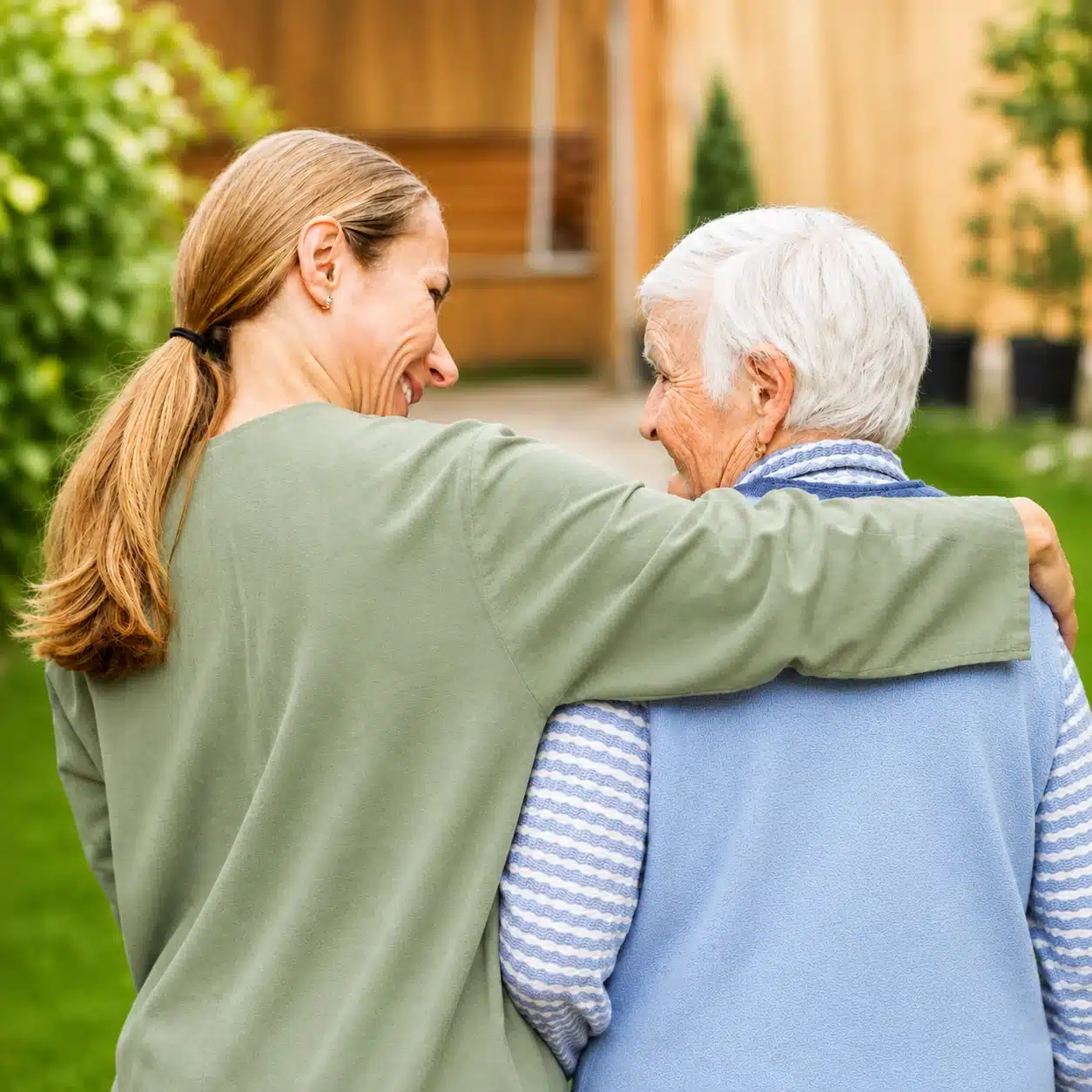 aphasia vs dementia A younger woman smiles and hugs an older woman as they walk outdoors, both seen from behind, enjoying a moment together in a garden or yard—a touching scene that highlights the warmth and connection possible even when loved ones are living with dementia.