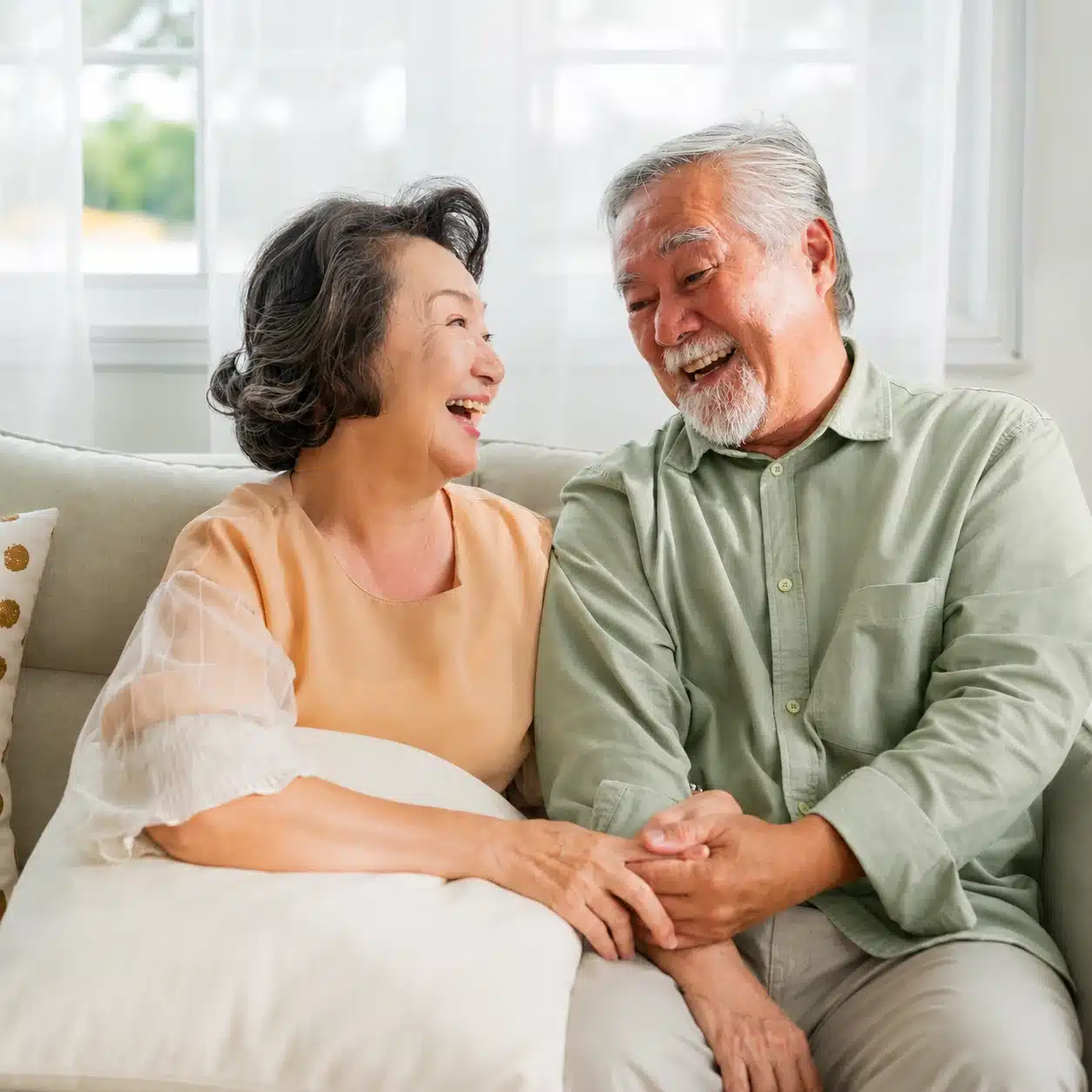 An elderly couple sits on a couch, smiling and laughing together while holding hands. Their joyful expressions reflect pure happiness as they enjoy each other’s company in a bright, cozy living room—an uplifting moment that also highlights the importance of aphasia awareness in fostering strong connections and communication.